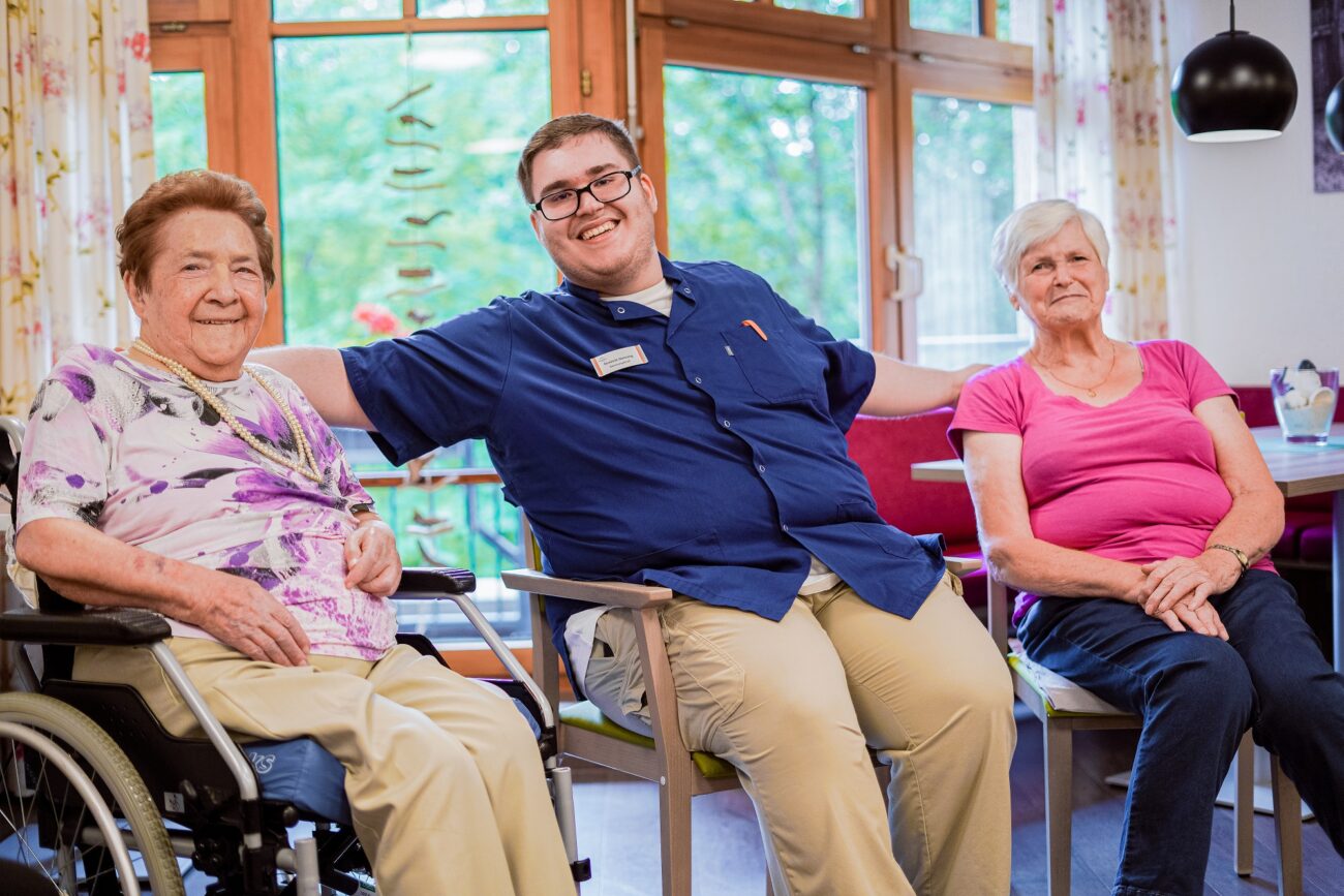 Herzlicher Betreuer im Bürgerheim Villingen-Schwenningen sitzt zwischen zwei Seniorinnen, eine im Rollstuhl, und führt ein freundliches Gespräch